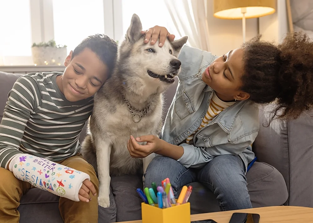 A boy with a broken arm and his sister are sitting on the couch and hugging their dog.